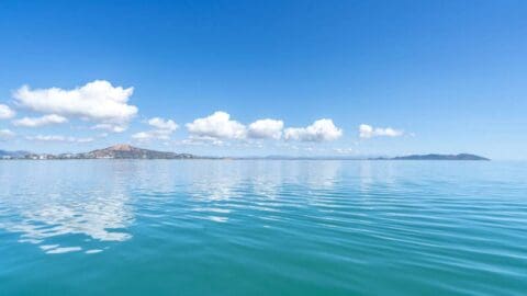 A calm blue ocean with small waves under a clear sky dotted with fluffy white clouds. A distant coastline with mountains is visible on the horizon.