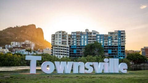 Large "Townsville" sign with background of modern buildings, trees, and a rocky hilltop at sunset.