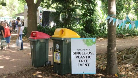 A sign reads "You are at a single-use plastic free event" next to recycling bins in an outdoor area with people and trees.