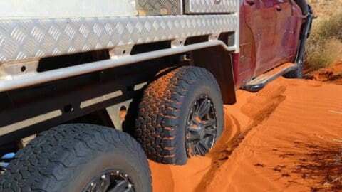 A red off-road vehicle with a metal utility body, perfect for ecotourism adventures, is stuck in deep red sand, with its rear tires partially buried.