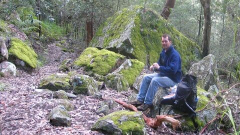 A man sits on a mossy rock in a forested area with a backpack nearby, taking a break on a sustainable hiking trail surrounded by trees and large moss-covered rocks.