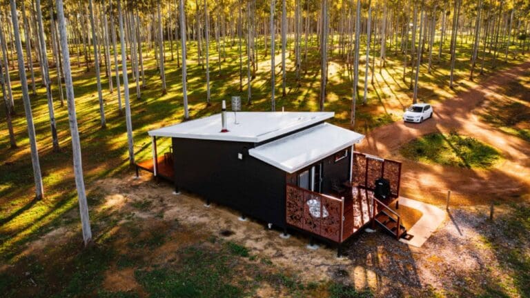 A small modern cabin with a white roof situated in a forest, surrounded by tall trees. Offering cozy accommodation for nature enthusiasts, the cabin is complemented by a white car parked on a dirt road next to it.