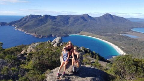 Three people sit on a rocky viewpoint overlooking a crescent-shaped turquoise bay surrounded by mountainous terrain and lush greenery under a clear blue sky, perfect for an ecotourism adventure.