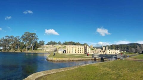 A wide-angle view of historic ruins surrounded by lush accommodation and water under a clear blue sky with a few clouds.