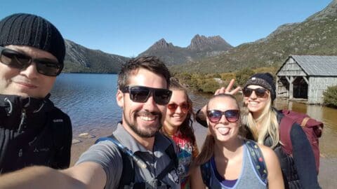 Five people pose for a selfie in front of a lake with mountains and a wooden hut in the background; two of them make playful gestures. This picturesque scene highlights their enjoyment at an eco-friendly accommodation.