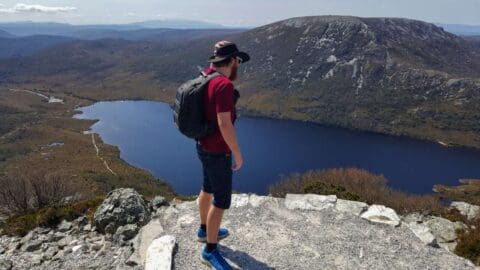 A person with a backpack stands on a rocky viewpoint overlooking a lake, with mountains and a valley in the background, as if embarking on an eco-tour.