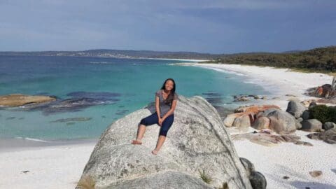 A person sits on a large rock at a beautiful beach with turquoise water, white sand, and rocky formations, under a clear sky, embracing the wonders of nature.