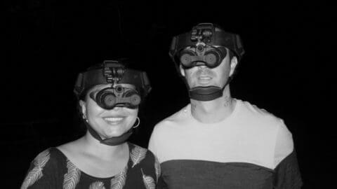 Two people, equipped with night vision goggles, stand together in the dark, smiling at the camera during an ecotourism adventure.