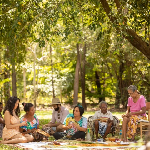 A group of people are seated on the grass under trees in a sunny outdoor setting, engaging in a sustainable crafting activity with natural materials.