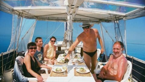 Five people are seated at a table on a boat. One person stands, pouring a drink. The weather is sunny, and the calm sea in the background enhances the serene connection with nature.