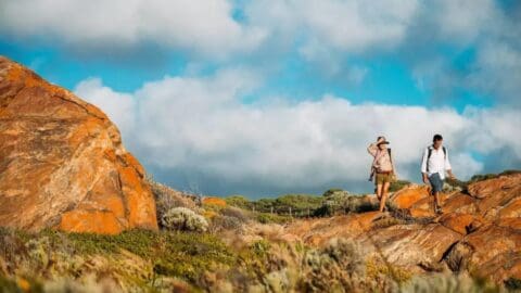 Two people hike on a rocky terrain with large, brown boulders and patches of greenery under a cloudy, blue sky, embracing ecotourism.