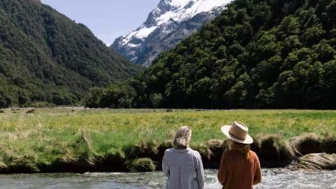 Two people, one wearing a hat, stand by a stream in a grassy valley surrounded by dense forest and snow-capped mountains, enjoying their sustainable tour.