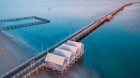 Aerial view of the Busselton Jetty extending over calm blue water with three small blue buildings on stilts, a boat moored in the distance, and a sectioned-off swimming area.