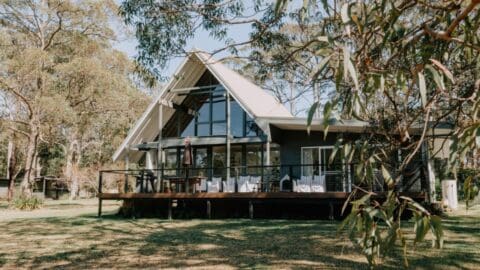 A modern, A-frame house with large glass windows and a spacious deck is surrounded by trees and greenery, offering sustainable accommodation ideal for ecotourism.
