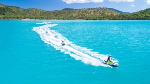 Several people are riding jet skis on a vibrant turquoise body of water near a hilly shoreline under a blue sky with scattered clouds. The area is known for its sustainable practices and offers eco-friendly accommodations for visitors.