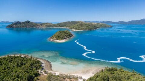 Aerial view of a tropical island surrounded by turquoise water and smaller islands, with a clear blue sky and white wake from a boat visible in the water, highlighting the beauty of unspoiled nature.