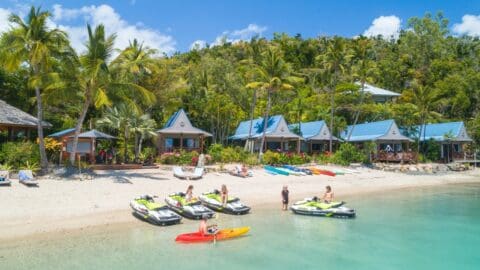 Tourists on a tropical beach with jet skis, kayaks, and beach houses surrounded by lush greenery, enjoying accommodation designed to blend seamlessly with nature.