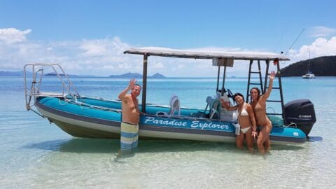Three people in swimwear stand and wave next to a "Paradise Explorer" tour boat in shallow, clear water, with a distant island and sailboats in the background.