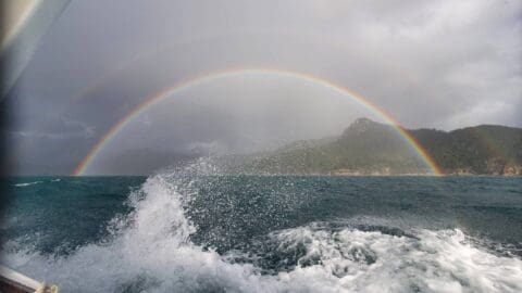 Experience the ocean waves with a rainbow in the sky amid cloudy weather, with mountainous land visible in the background. Join our sustainable tour for an unforgettable adventure.