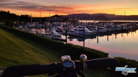 A view of a marina at sunset with numerous docked boats and mountains in the background, seen from behind handlebars during an ecotourism tour.