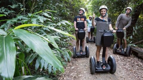 Four people riding Segways along a forest trail, surrounded by dense greenery. All are wearing helmets and casual clothing. The woman at the front is giving a thumbs-up, making the nature tour even more enjoyable.