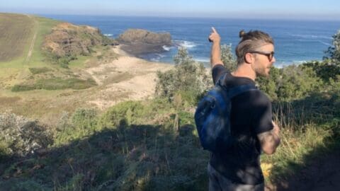 A man with a backpack points towards a coastal landscape with cliffs and the ocean in the background, while standing on a hill covered in vegetation during an eco-friendly tour.