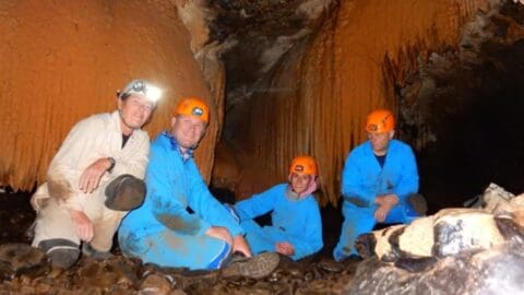 Four people wearing helmets and blue overalls are sitting inside a cave with stalactites and stalagmites visible in the background. They appear to be certified caving explorers, showcasing the wonders of ecotourism.