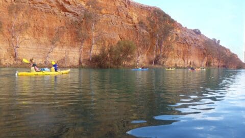 A group of people are kayaking on a calm river next to a tall, rocky cliff with sparse vegetation, enjoying the beauty of nature.