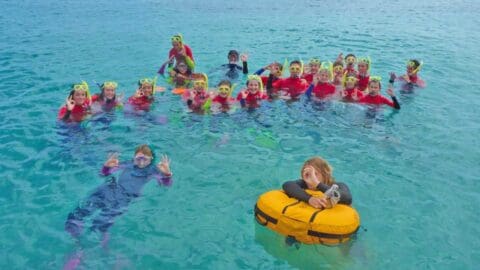 A group of people in snorkeling gear wave and smile while floating in clear blue water, with one person resting on an inflatable yellow ring at the front, enjoying their tour.