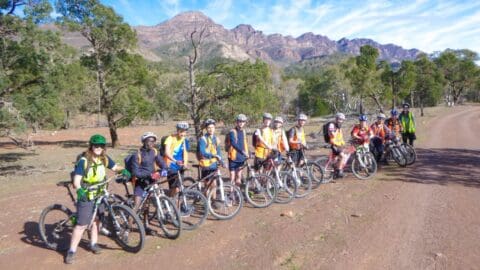 A group of cyclists, sporting helmets and orange vests, pose with their bikes on a dirt path in a scenic, mountainous area with trees. They are part of an organized tour known for its excellent accommodation options and professional certification for guides.