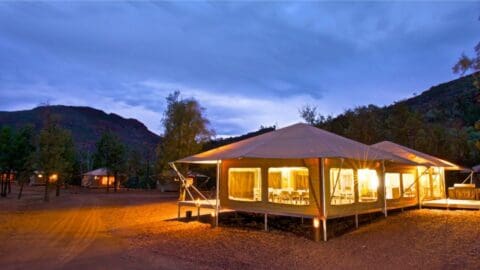 A large, lit-up tent sits on a platform in a desert area at dusk with mountains in the background, offering sustainable accommodation for ecotourism enthusiasts.