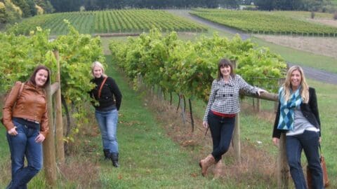 Four women stand and pose in a vineyard. The rows of grapevines stretch into the distance, with a variety of greenery and fields in the background. They are dressed casually and smiling, perhaps taking a break from their adventure with Simmo's Offroad Tours.