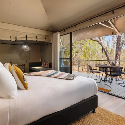 A bedroom with a large bed, colorful pillows, and blanket. A glass sliding door leads to a patio with outdoor furniture, overlooking a serene wooded area. This accommodation offers the perfect escape for nature enthusiasts.