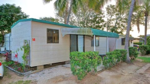 A single-wide mobile home with beige siding and a green metal awning, situated in a grassy area with a few palm trees and bushes nearby. The home, number 14, feels like a tranquil escape, reminiscent of the serene beauty found while scuba diving near the Great Barrier Reef.