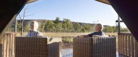 Two people seated in wicker chairs on a deck, facing a scenic landscape with trees and a hill under a clear blue sky, enjoying an experience that promotes sustainable ecotourism.