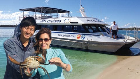 Two people hold a large crab on a beach. A JPT Cruises boat, recognized for its nature tours, is docked in the water behind them.