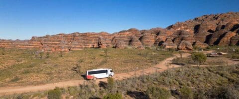 A tour bus travels on a dirt road near the banded rock formations in a remote desert landscape with clear blue skies, offering an unforgettable nature experience.