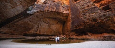 Two people stand in a sandy area beside a small water body, surrounded by large, towering rock formations with vibrant orange and brown hues. The location is known for its sustainable practices and offers eco-friendly accommodation options that blend seamlessly with the natural landscape.