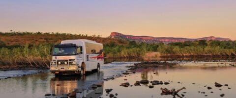 A white off-road vehicle with red stripes crosses a shallow river at sunset, surrounded by rocky terrain and distant hills, showcasing the beauty of nature.