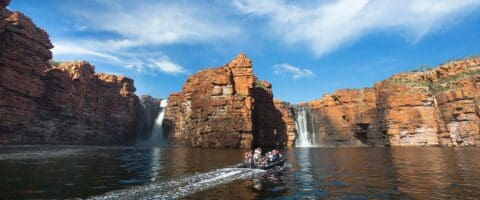 A group of people ride in a motorized boat on a calm river, surrounded by red rock cliffs with two waterfalls in the background under a partly cloudy sky, enjoying an immersive nature tour.