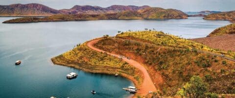 A winding road leads to a boat ramp on a small peninsula surrounded by a large lake with hilly terrain in the background. Several cars and boats are present near the shoreline, perfect for those seeking nature tours or lakeside accommodation.