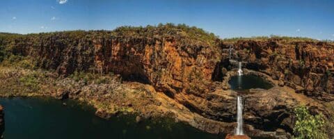 A panoramic view of a rocky canyon with a cascading waterfall flowing into multiple ponds below under a clear blue sky. Sparse vegetation covers the canyon top, showcasing the raw beauty of nature and making it an ideal spot for ecotourism.