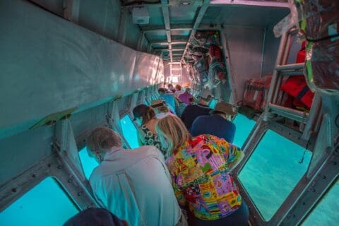 People in colorful clothing lean forward to look out the windows of a submarine, observing the underwater views through the glass, offering a unique ecotourism experience.