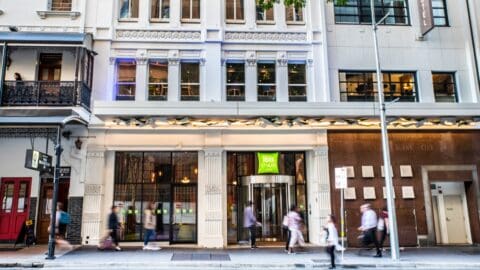 Street view of an Ibis Styles hotel entrance with pedestrians walking by. The building features tall windows, decorative trim, and a canopy above the entrance, showcasing a modern accommodation option.