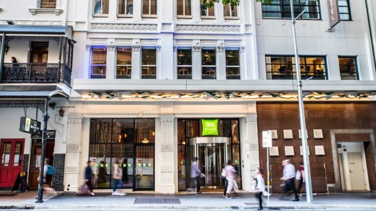 Street view of an Ibis Styles hotel entrance with pedestrians walking by. The building features tall windows, decorative trim, and a canopy above the entrance, showcasing a modern accommodation option.