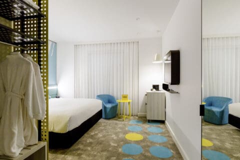 A modern ibis Styles hotel room in East Perth with a bed, two blue chairs, a TV, a yellow side table, and a hanging bathrobe. The room features a minimalist design with polka dot carpet accents.