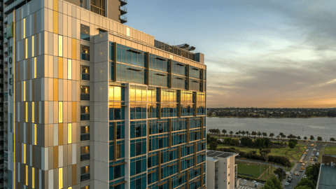 Modern high-rise building with glass facade reflecting a sunset, overlooking a waterfront with trees and a road lined with cars, near the vibrant ibis Styles in East Perth.