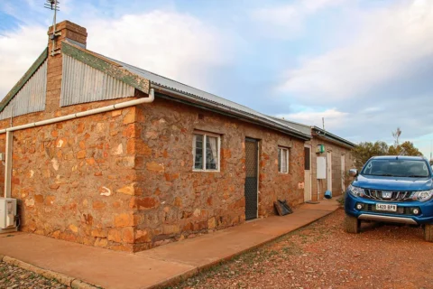 A stone house with a metal roof, nestled in the rugged terrain of Bendleby Ranges. A blue pickup truck is parked beside it. The ground is a mix of concrete and gravel, and the sky is partly cloudy.