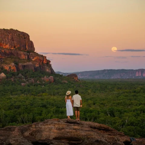 A couple stands on a rocky outcrop, overlooking a forested landscape with a cliff on the left. The sun is setting, casting a warm glow across the scenery, perfect for an ecotourism tour.