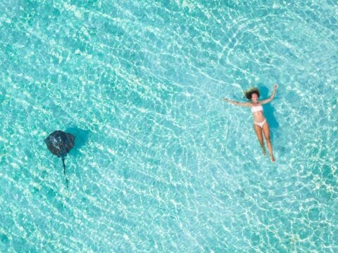 A woman floats on her back in clear, turquoise water while a stingray swims nearby, showcasing the beauty of sustainable ecotourism.